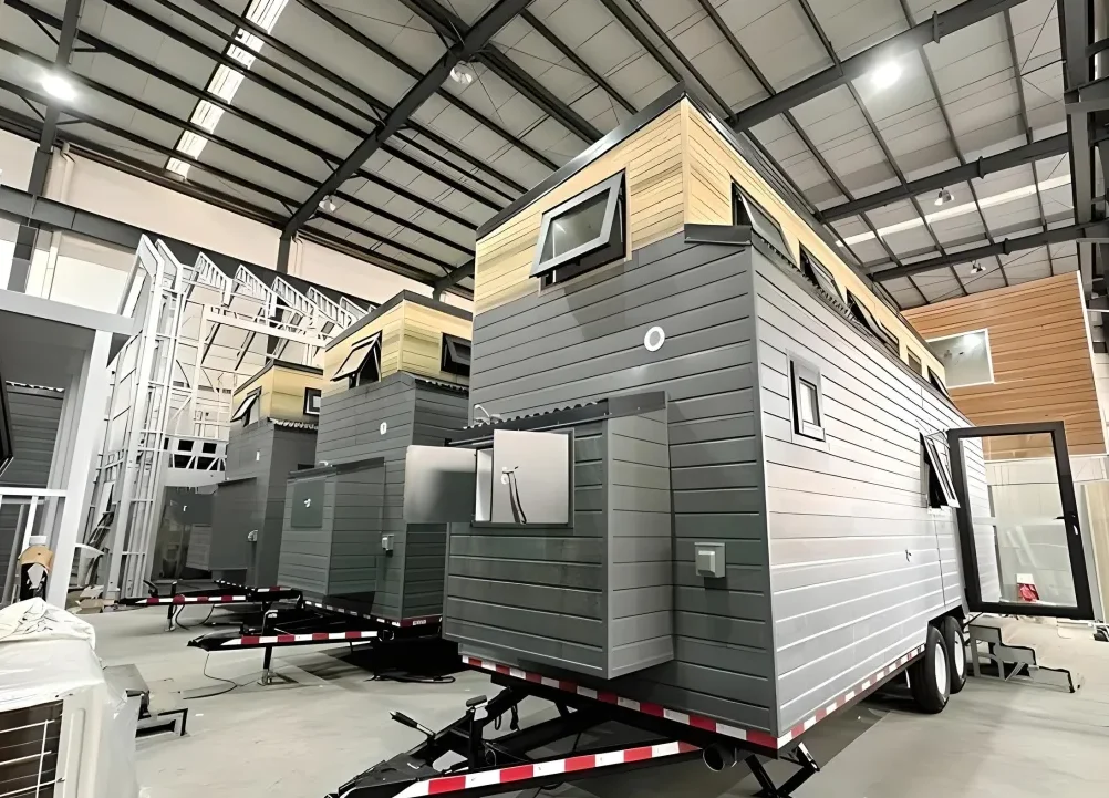 Tiny homes part built on trailers inside a factory workshop, grey and timber cladding, with window frames stacked behind them
