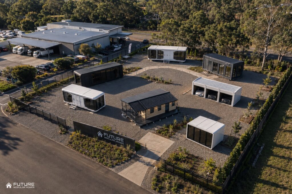 Aerial view of a display village with capsule homes, a verandah cabin and a three bay garage laid out on gravel beside a warehouse