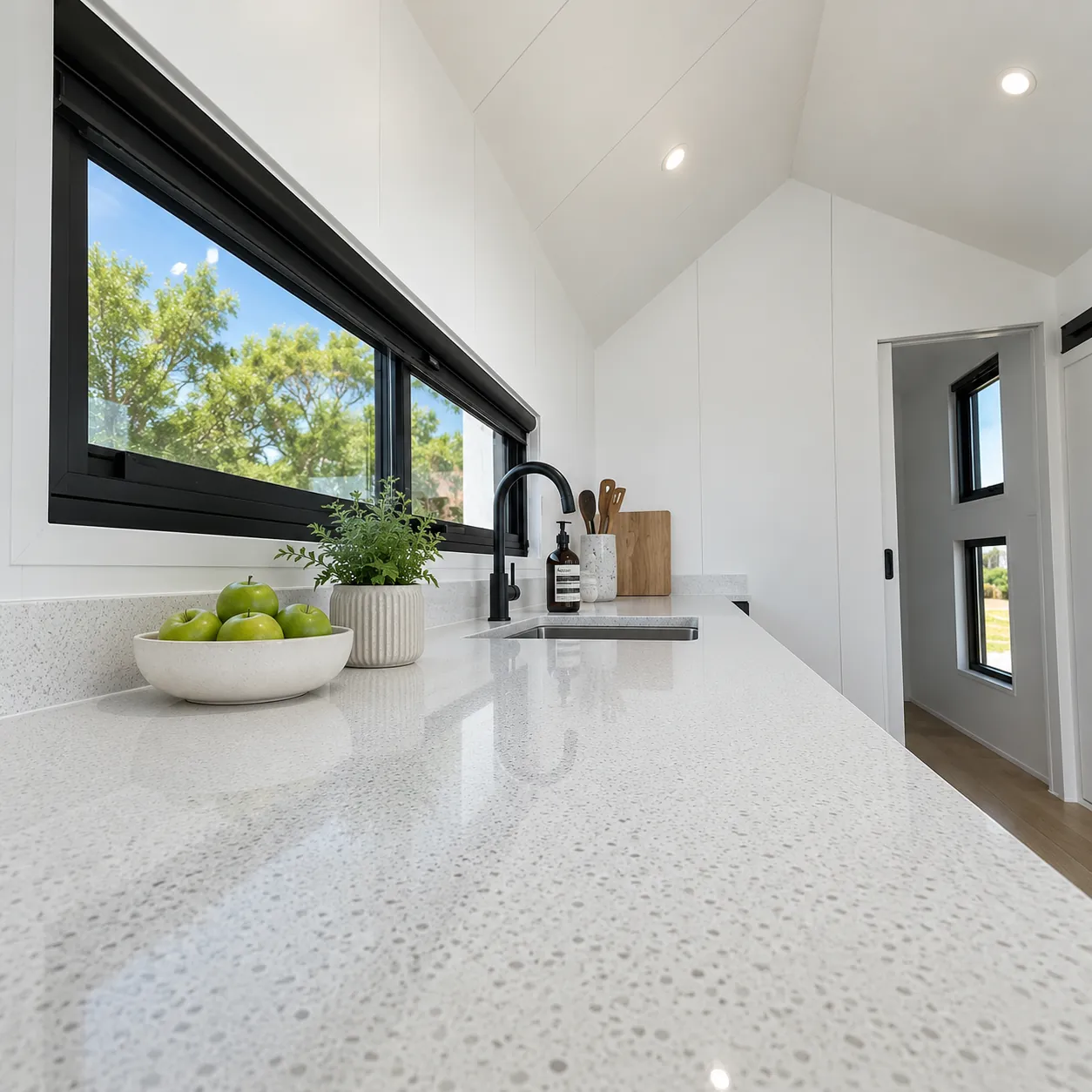 Galley kitchen in a tiny home, white speckled benchtop with an undermount sink and a black gooseneck mixer, a long black framed window above the bench and a raked white ceiling with downlights