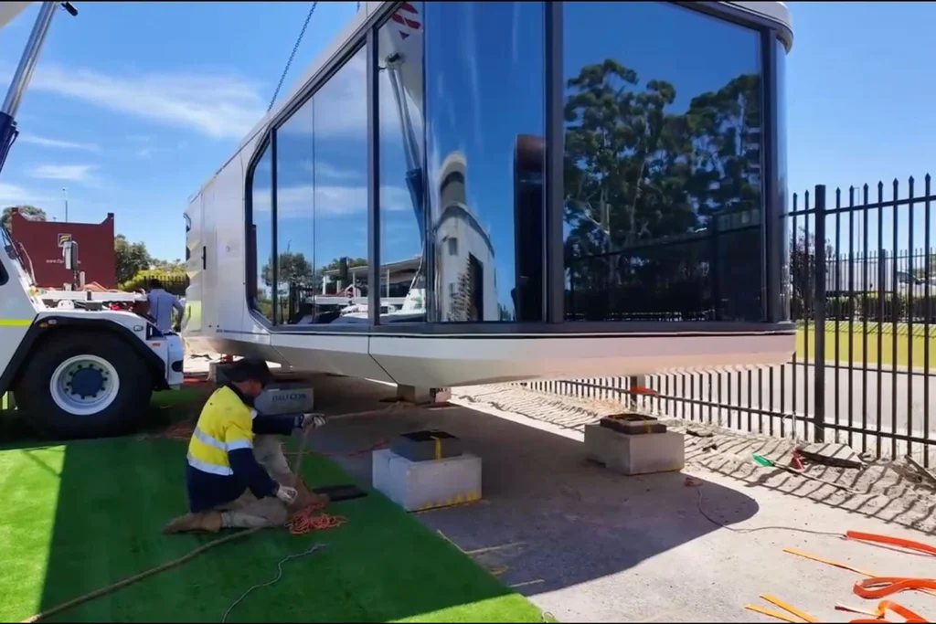 Worker in a hi-vis shirt kneeling with rigging rope beside a capsule home as a crane lowers it onto concrete pier blocks