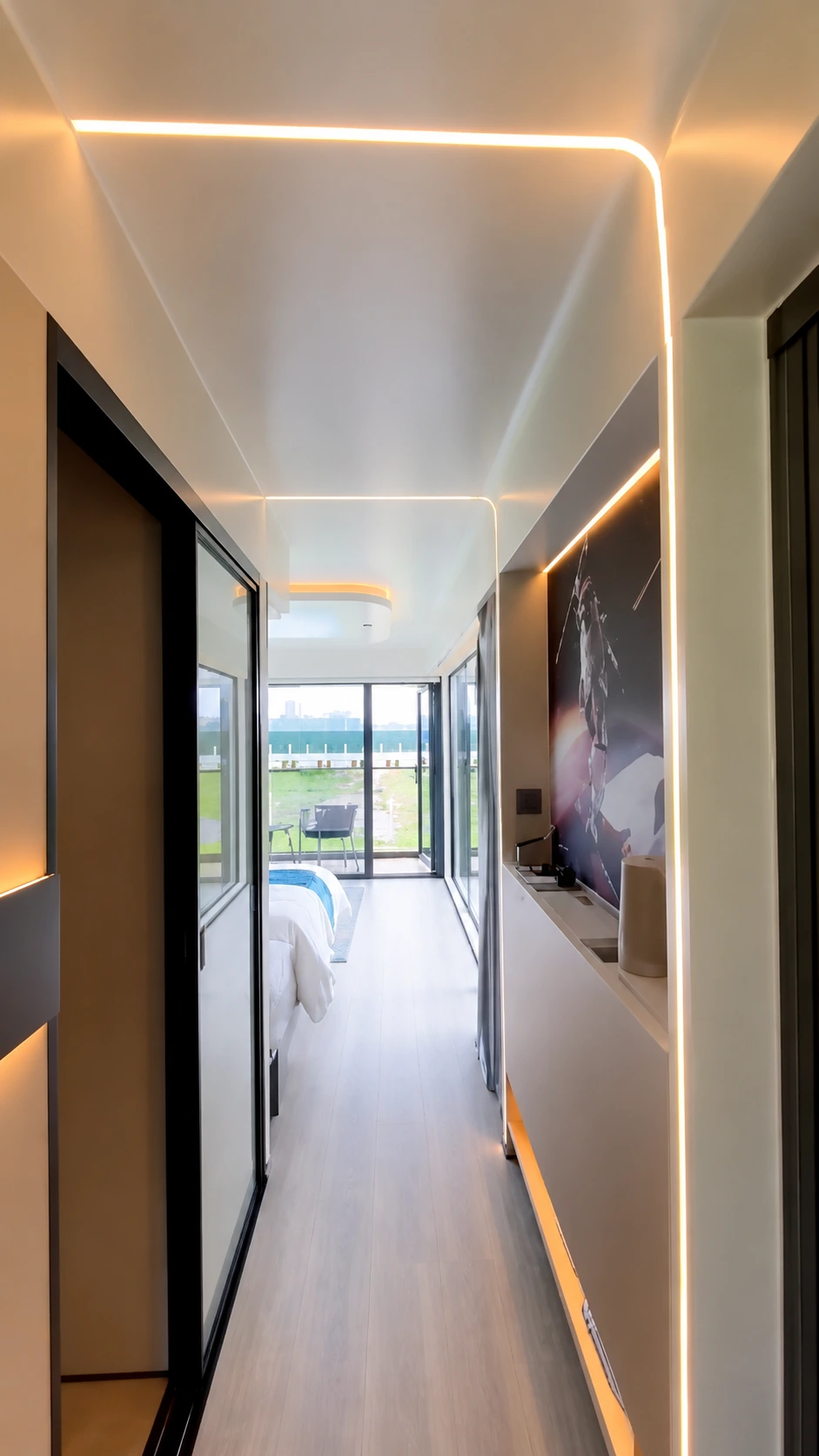 Internal corridor of a prefab home with timber look flooring, a curved LED strip running across the ceiling, a frosted glass sliding door to the bedroom on one side and glazed doors at the end opening onto a deck