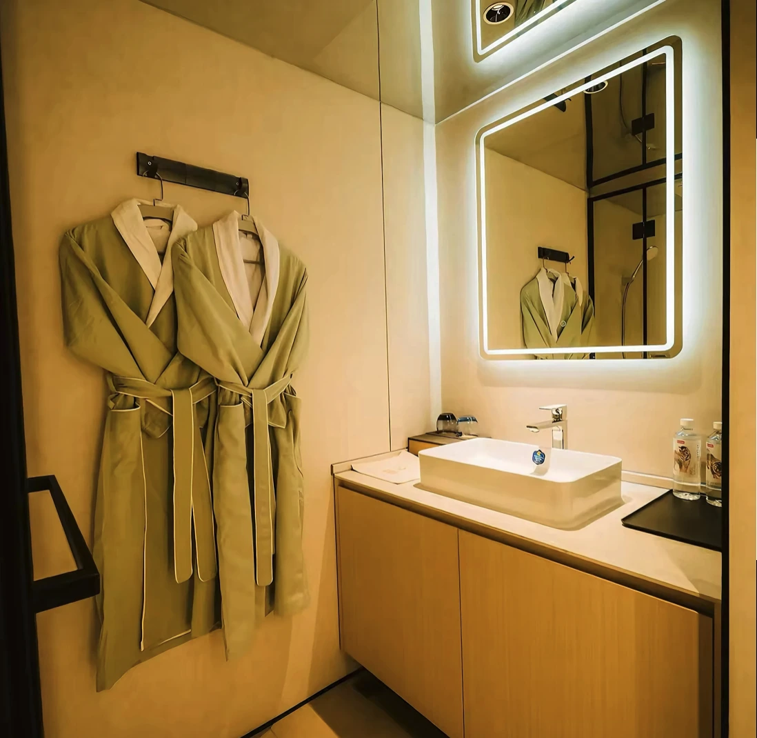 Compact bathroom with two robes hanging on a black wall rail, a backlit square mirror above a white vessel basin on a timber vanity, and a black framed shower screen reflected in the mirror