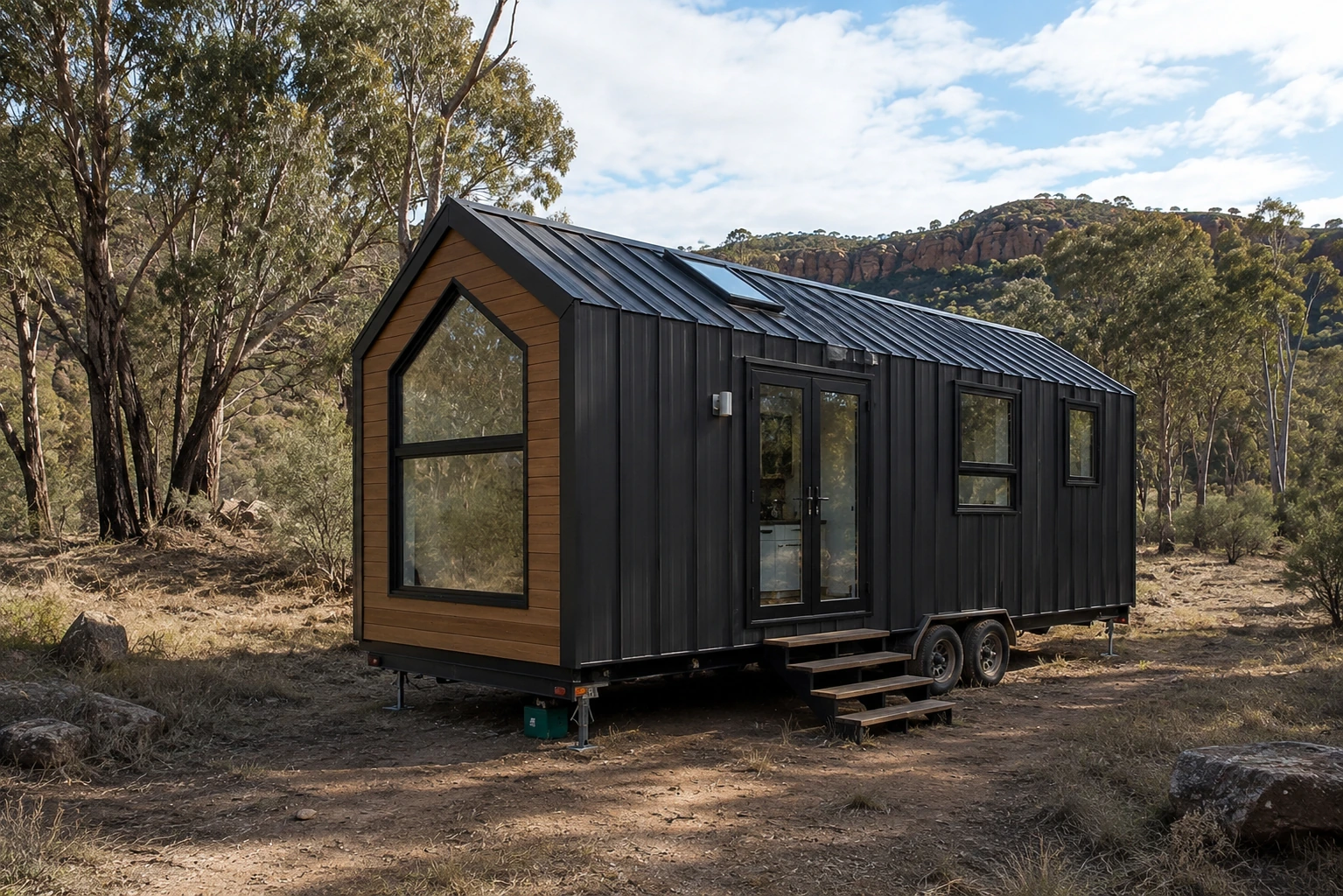 Black tiny house on a tandem axle trailer standing on levelling jacks, with a timber clad gable end, a tall arched window, glazed double doors and timber steps, on a dirt clearing in eucalypt bushland