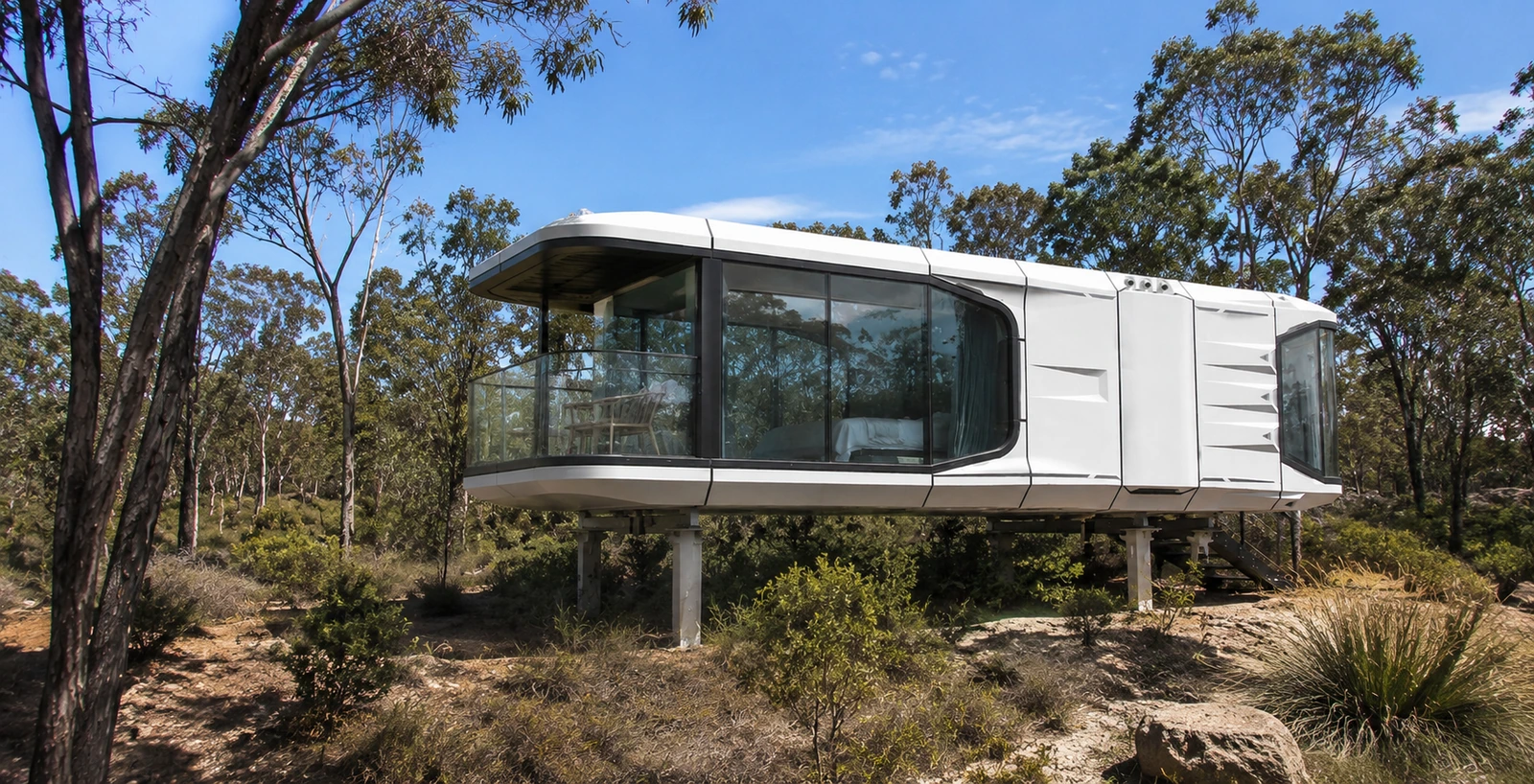 White capsule home raised on slender columns above sloping bushland, with wraparound tinted glazing and a glass balustrade deck, gum trees and grass trees around it