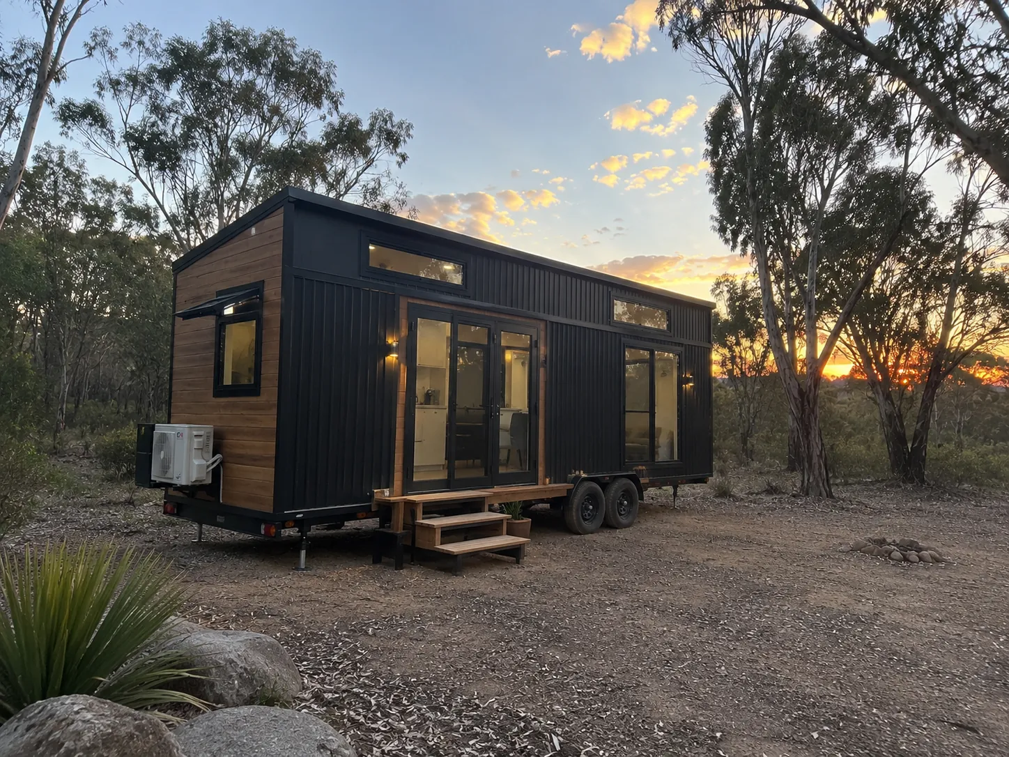 Tiny home on a tandem axle trailer with black corrugated and timber cladding, lit glazed doors, timber entry steps and a split system condenser on the end wall, parked on gravel among gum trees at sunset