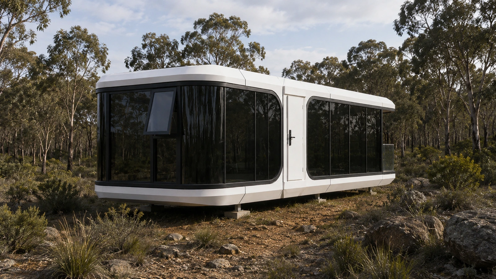 A white modular capsule home with black glazing sitting on low concrete pier footings in open Australian bushland, surrounded by gum trees, rocky ground and scrub