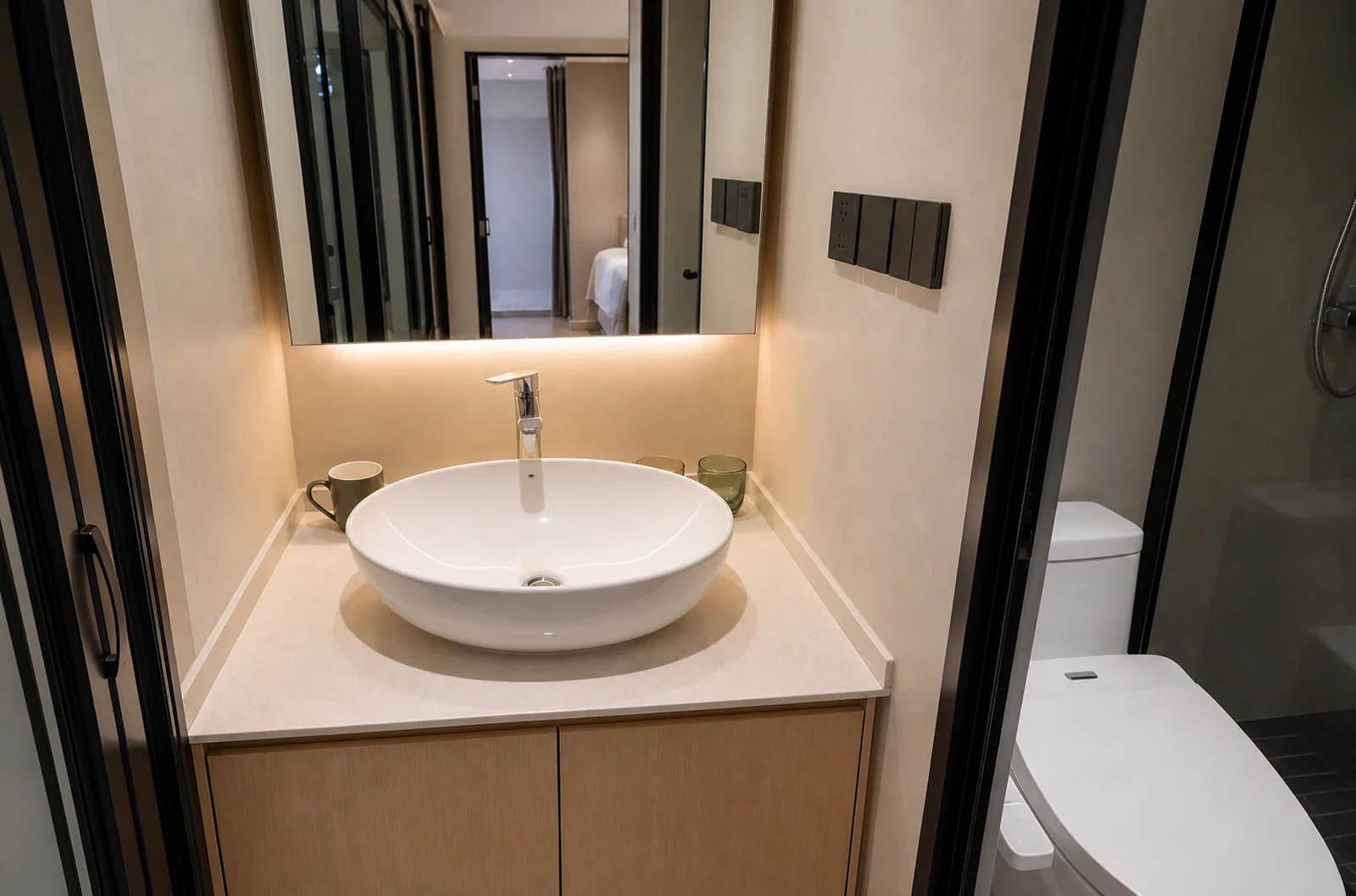 Bathroom in the compact capsule, round basin on a timber vanity under a lit mirror with the toilet beside it and the bedroom reflected behind