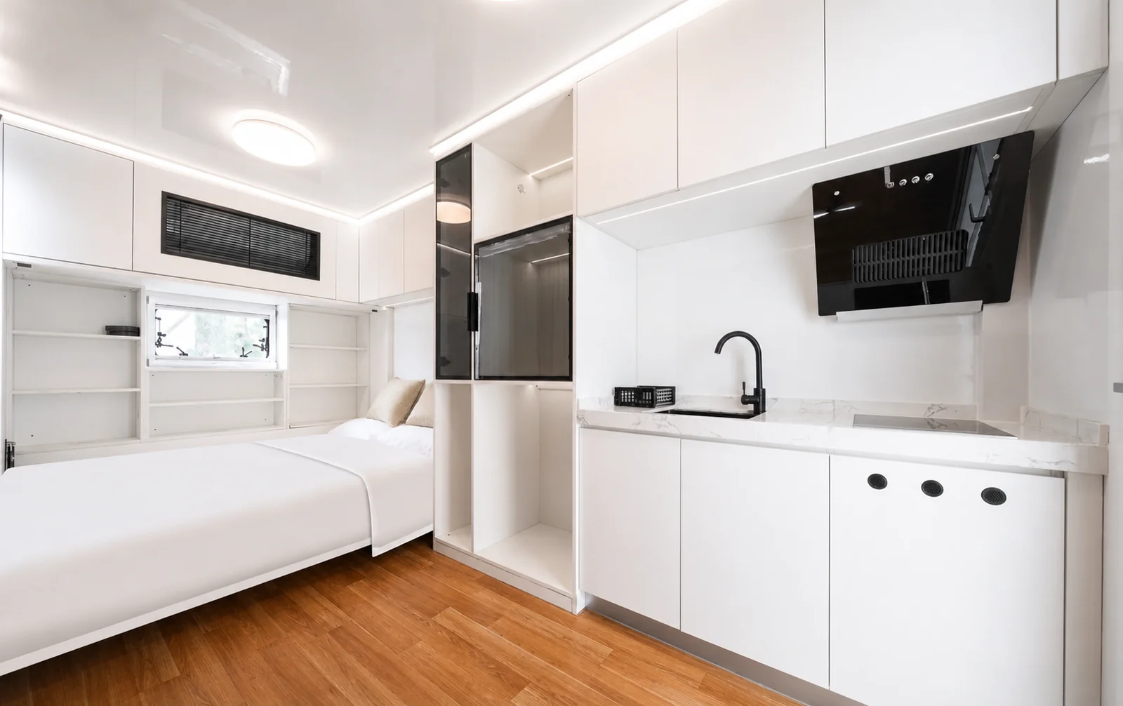 Interior of the 5.8 m home, double bed to the left of a full height storage wall and a white kitchen run with sink and cooktop on the right, oak floor