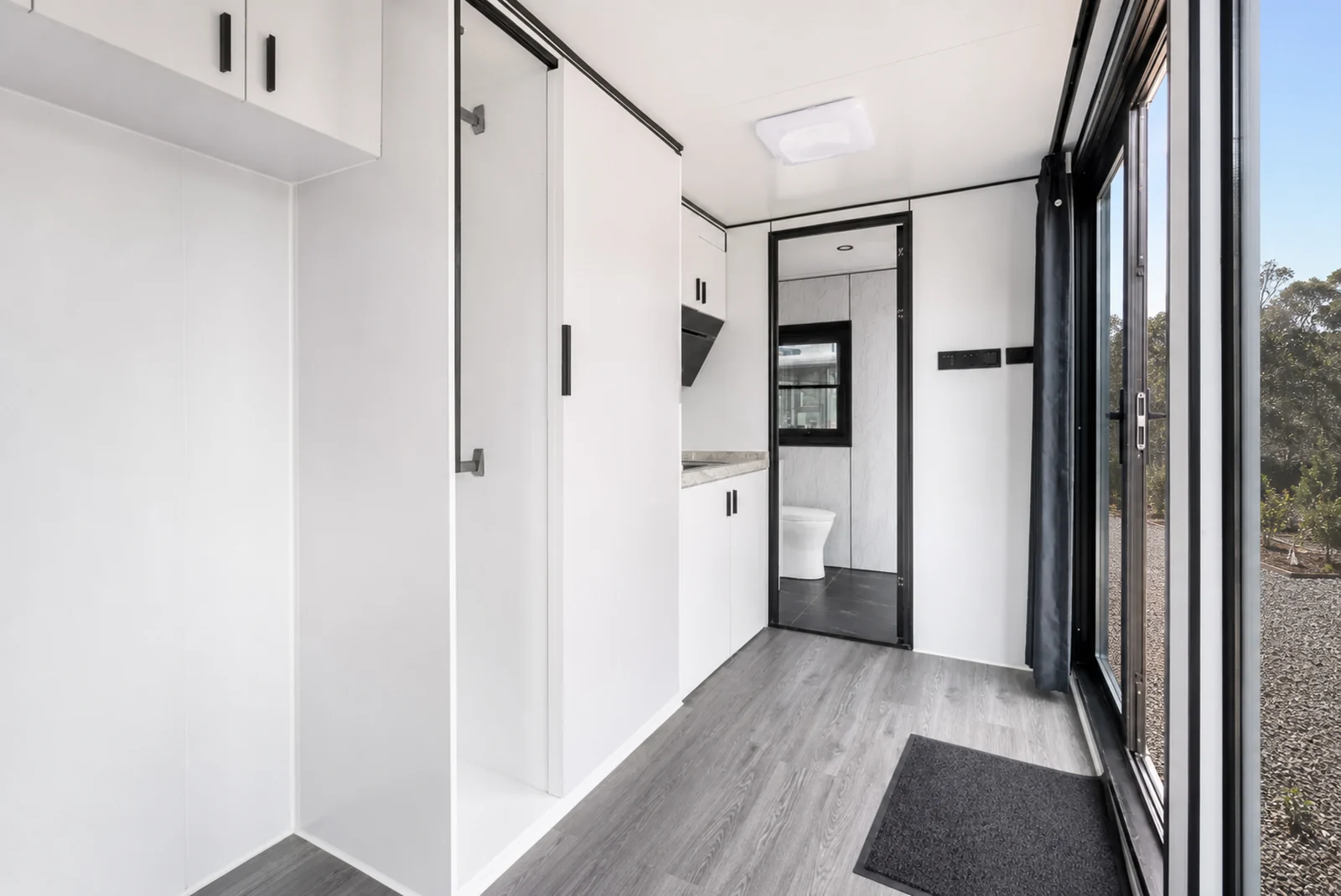 Looking down the 5.8 m home from the entry, storage cupboards on the left and the bathroom door open to show the toilet, grey timber look vinyl floor