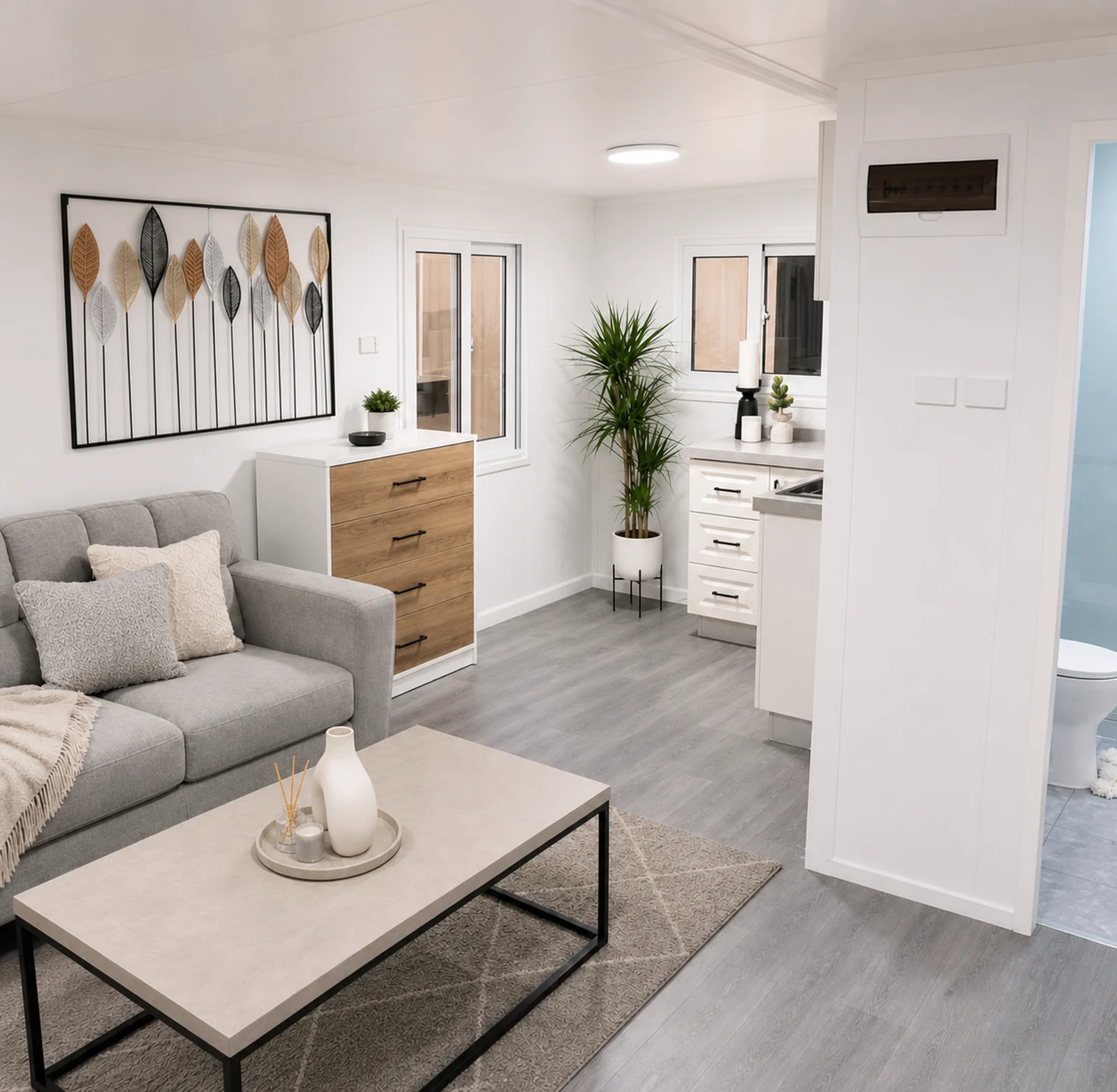 Living area inside the expandable home, grey sofa and coffee table on a rug with a timber drawer unit and the bathroom door open beyond