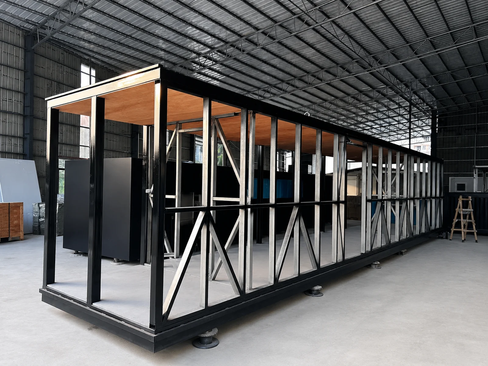 The 11.5 m mirror house as a bare galvanised steel frame in the factory, braced wall studs and black perimeter beams standing on adjustable levelling feet before the mirror glass is fitted