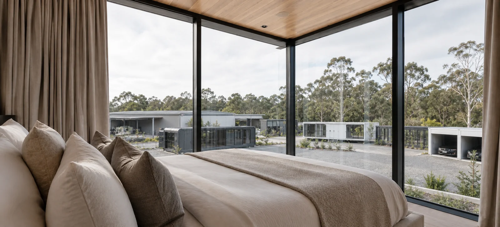 Bedroom in the wide mirror house, made bed in linen against a full height glazed corner with a timber lined ceiling and heavy curtains