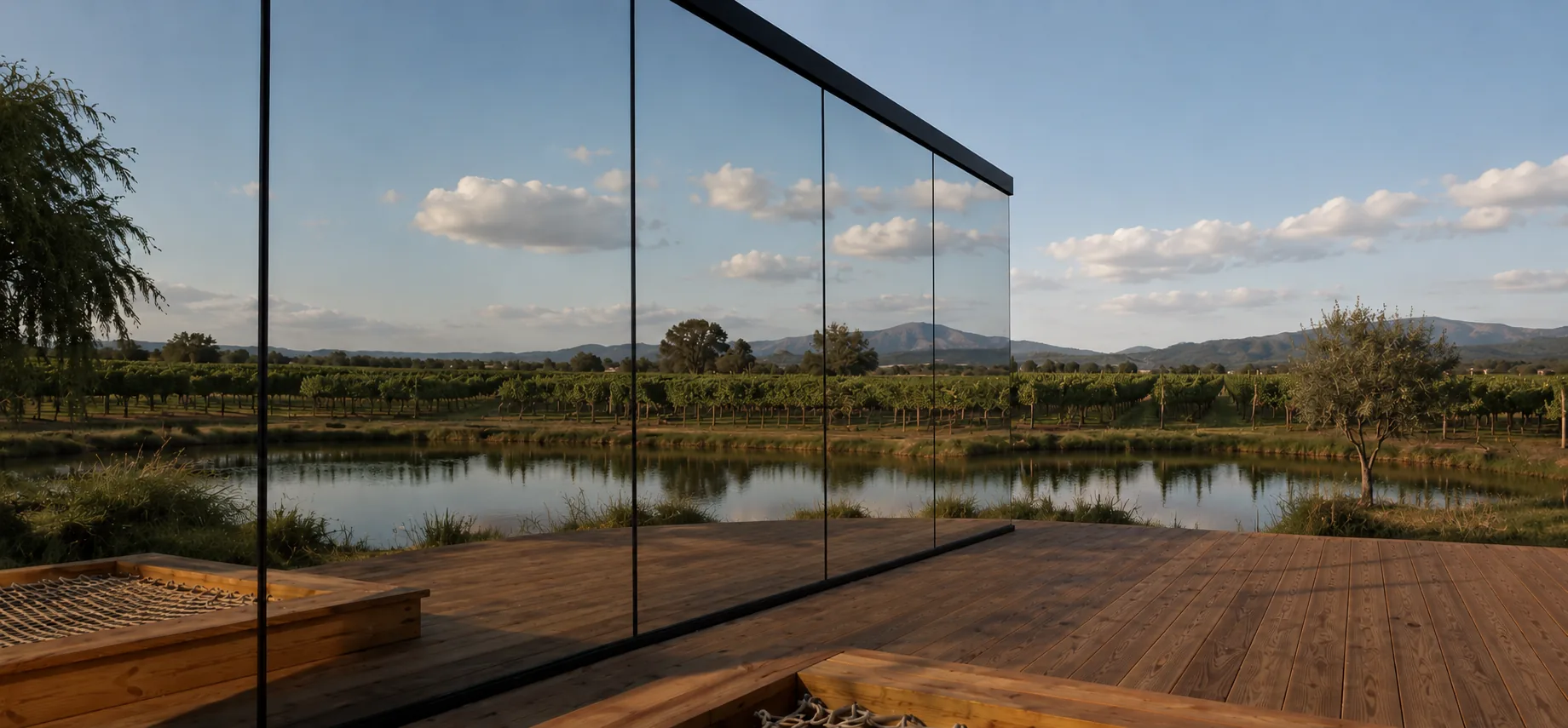 The 8 m mirror house beside a timber deck, its mirrored glass reflecting a dam, rows of vines, low hills and a cloudy sky
