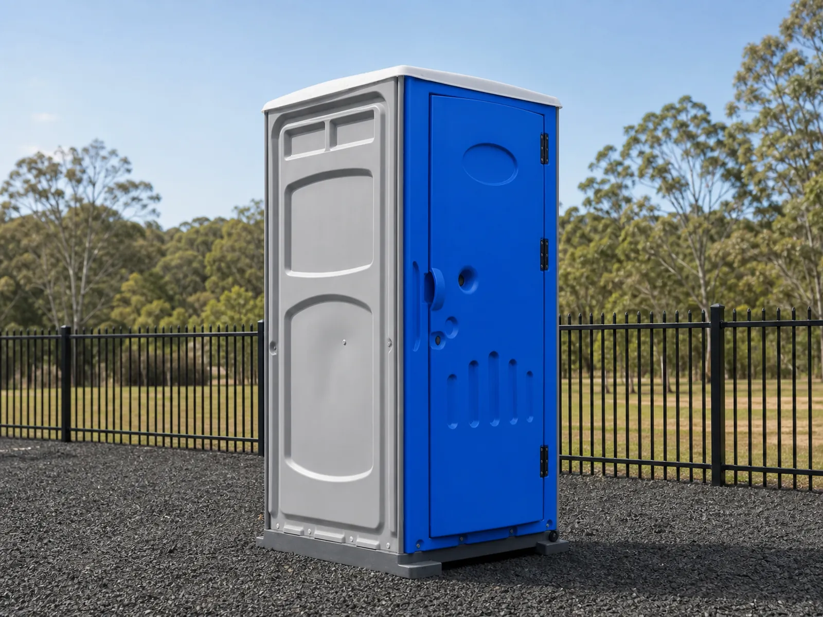 Blue and grey portable toilet standing on gravel beside a black steel fence with gum trees behind it