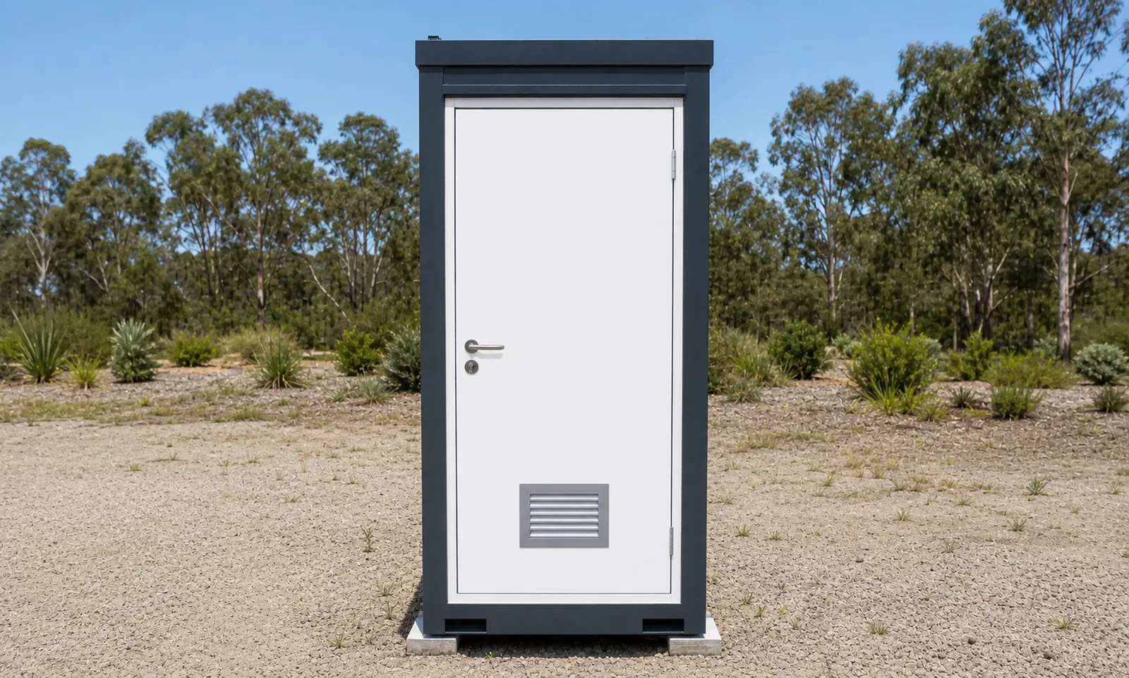 Narrow steel toilet cabin with a charcoal frame and a white door with a lever handle and a louvre vent, standing on levelling pads in dry bush