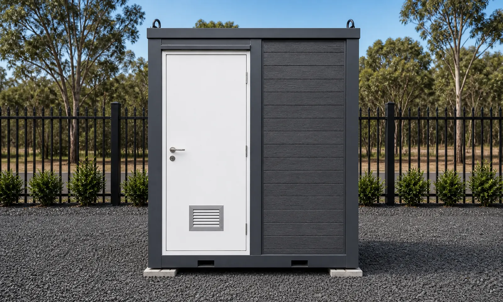 The portable bathroom unit outside on a gravel pad, white entry door with a floor vent and timber grain cladding on the side wall
