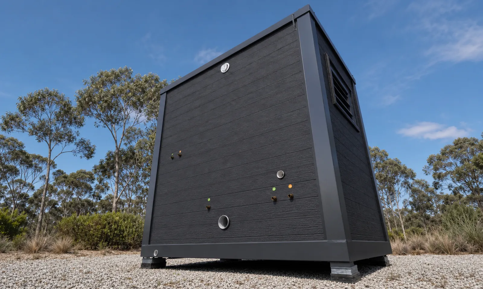 The portable bathroom from below the corner, dark timber grain cladding with service inlets and a louvre vent, standing on adjustable feet on gravel