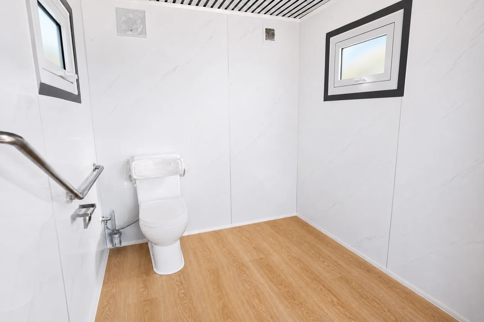 Inside the accessible toilet, toilet against the wall with a fold down grab rail and hand spray, two windows and timber look vinyl flooring