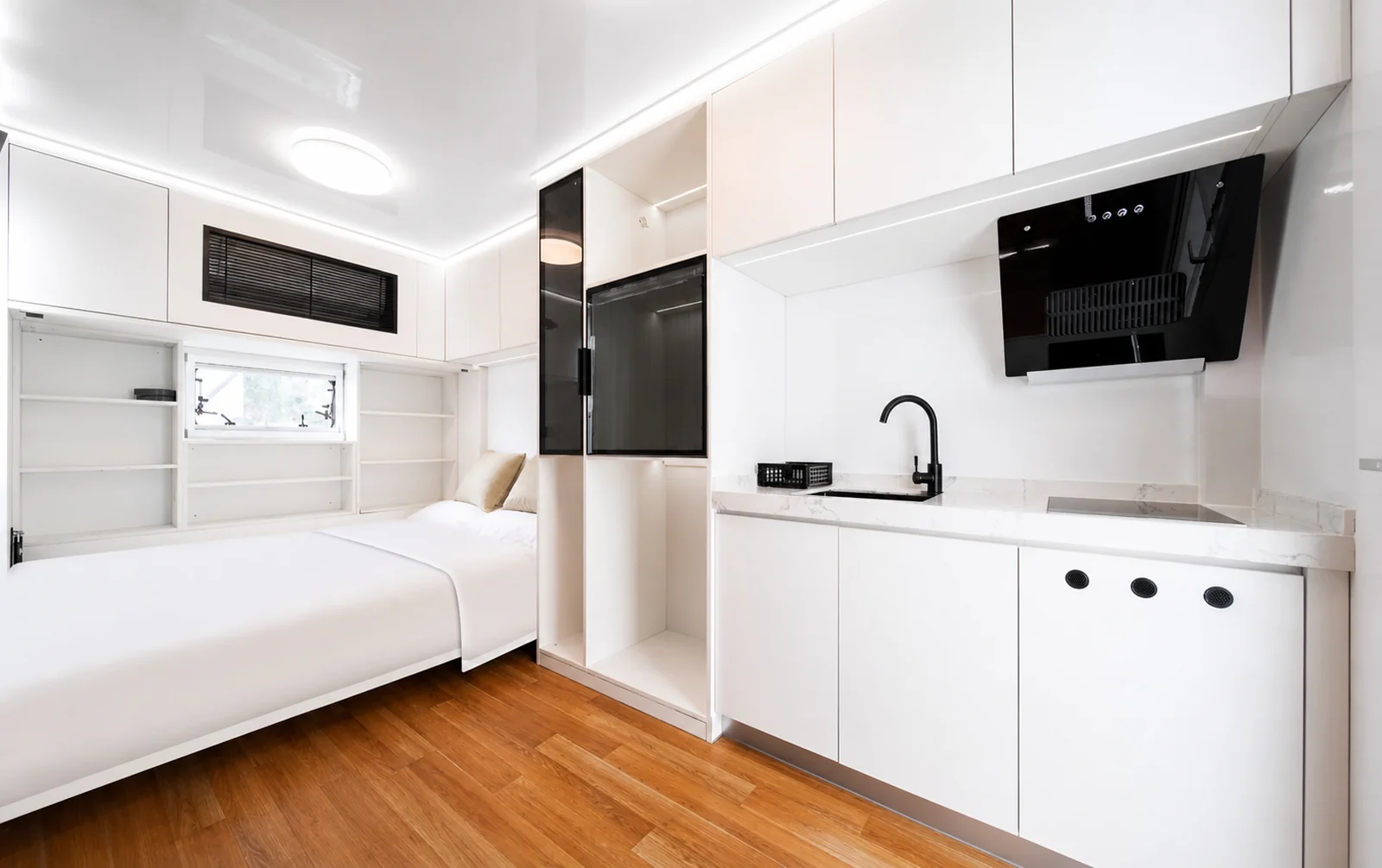 Inside the rounded capsule, single bed against the far wall beside a white kitchen run with a rangehood, sink and black mixer