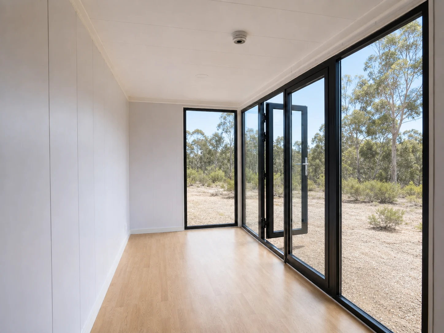 Inside the empty 6 m office, white lined walls and timber look vinyl floor with the glazed corner doors open to the bush