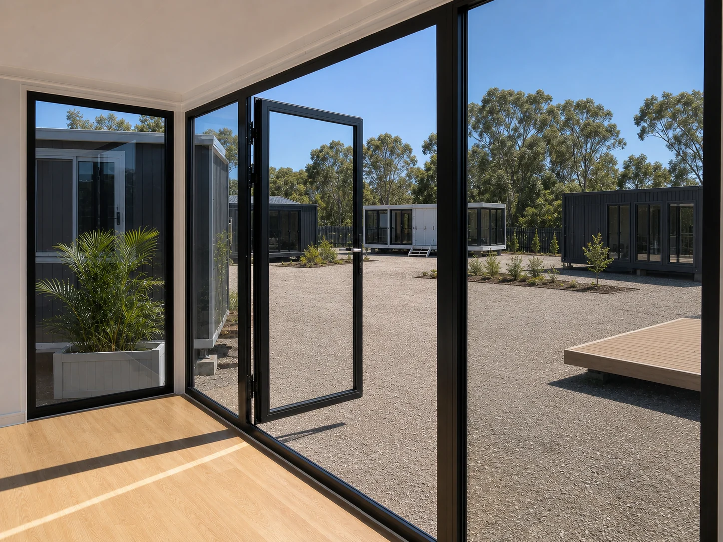 Looking out of the 6 m office through the open corner doors to a gravel yard with other cabins and a small timber deck