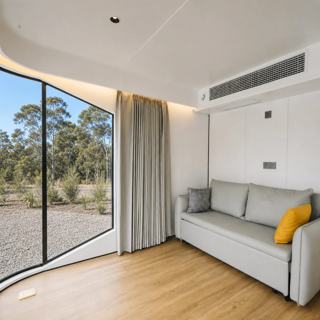 Living end of the capsule studio, pale grey sofa with a mustard cushion against the curved white wall beside a full height curtained window