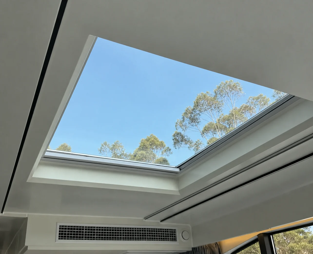 Looking up at the laminated glass skylight in the capsule roof with gum trees and blue sky above and a ceiling air outlet below