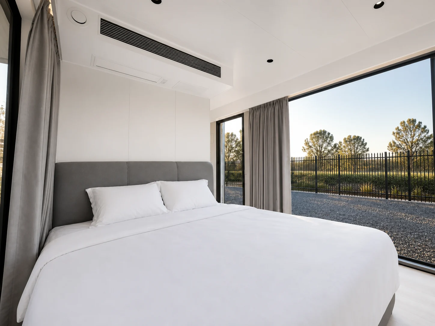 Bedroom in the balcony capsule with the curtains open, white bedding, a padded bedhead and full height glazing to the gravel pad