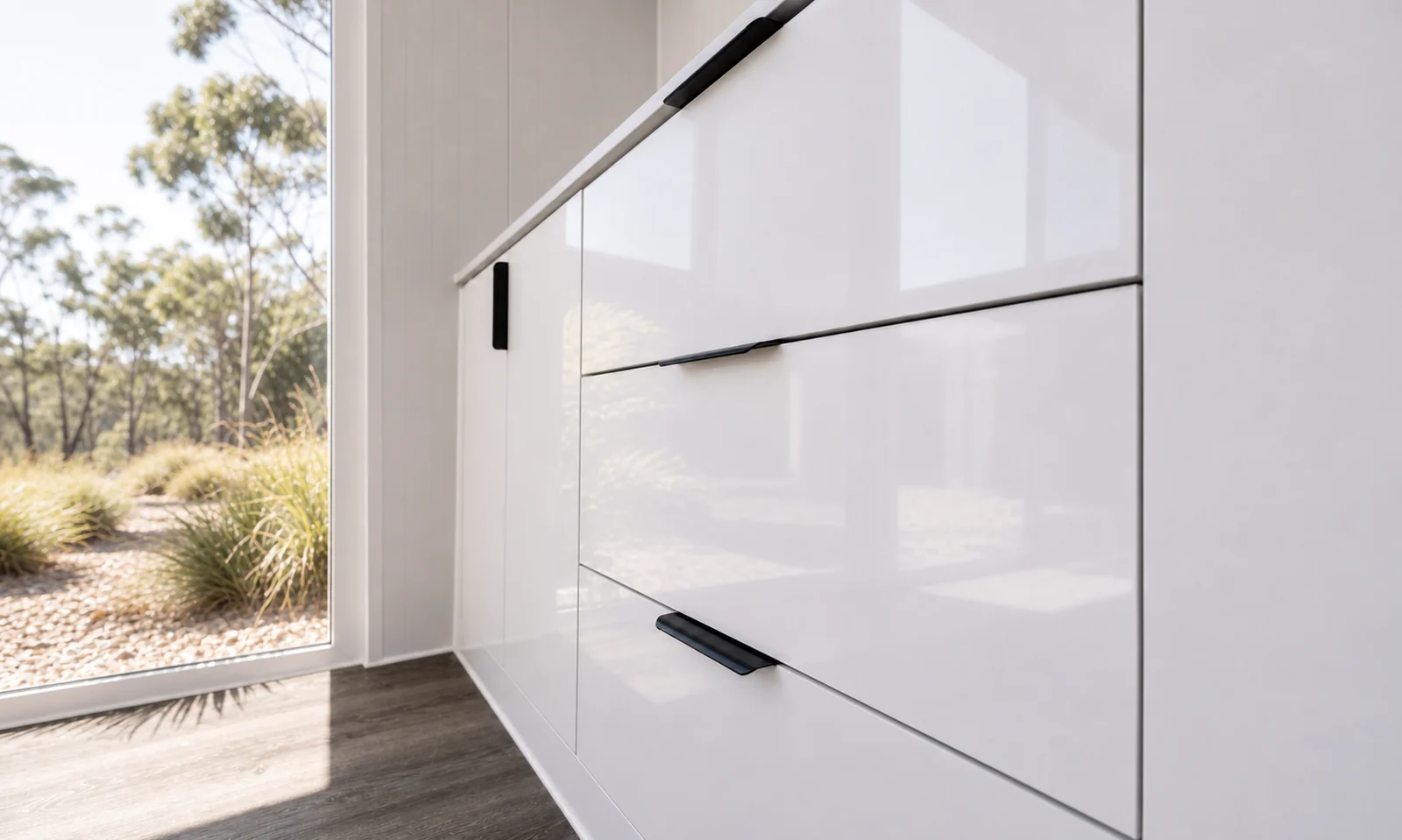 Close view of the gloss white drawer fronts and black pull handles beside the glazed door with the bush outside