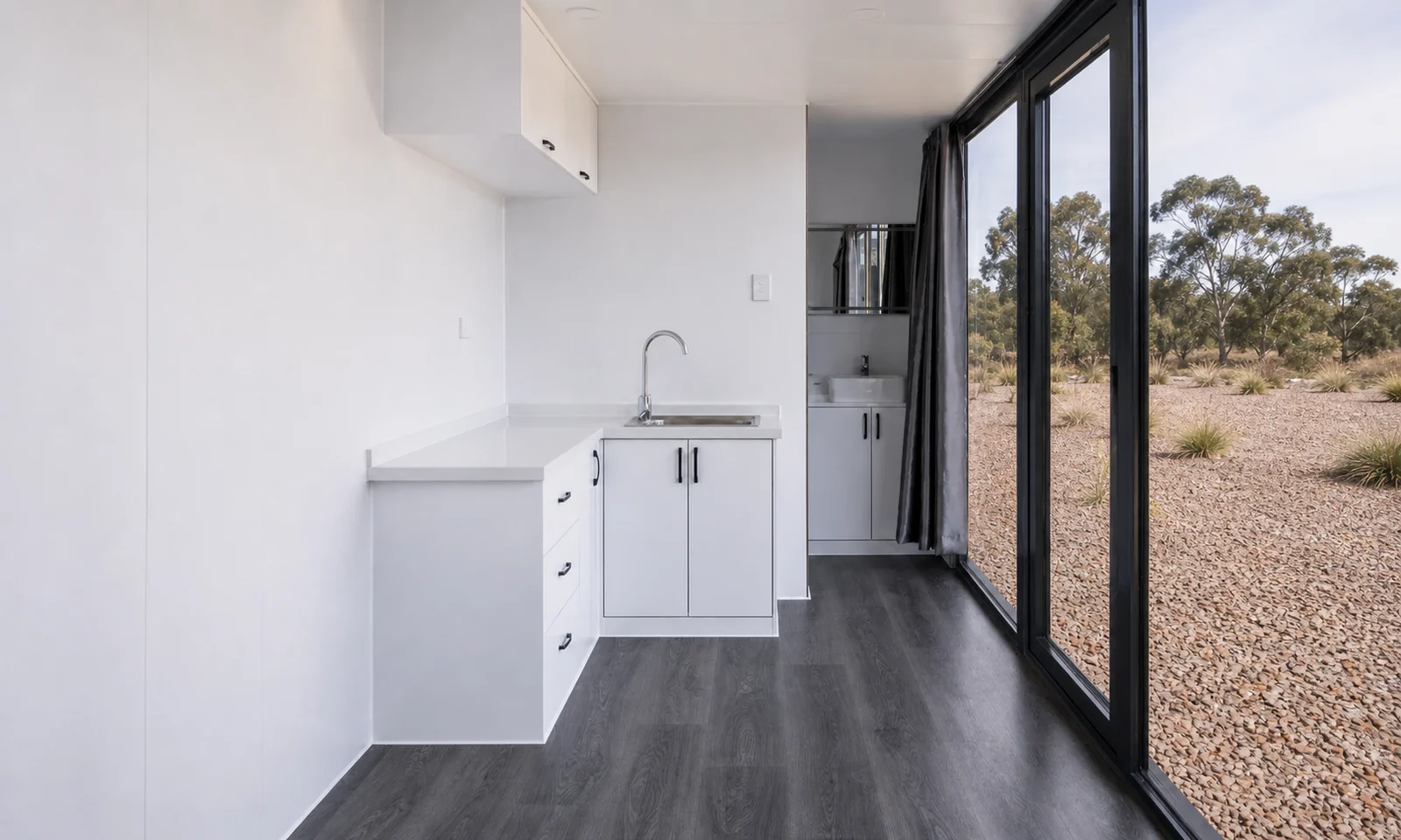 Kitchenette inside the studio cabin, white cabinets with drawers, a stainless sink and overhead cupboard beside the glazed wall
