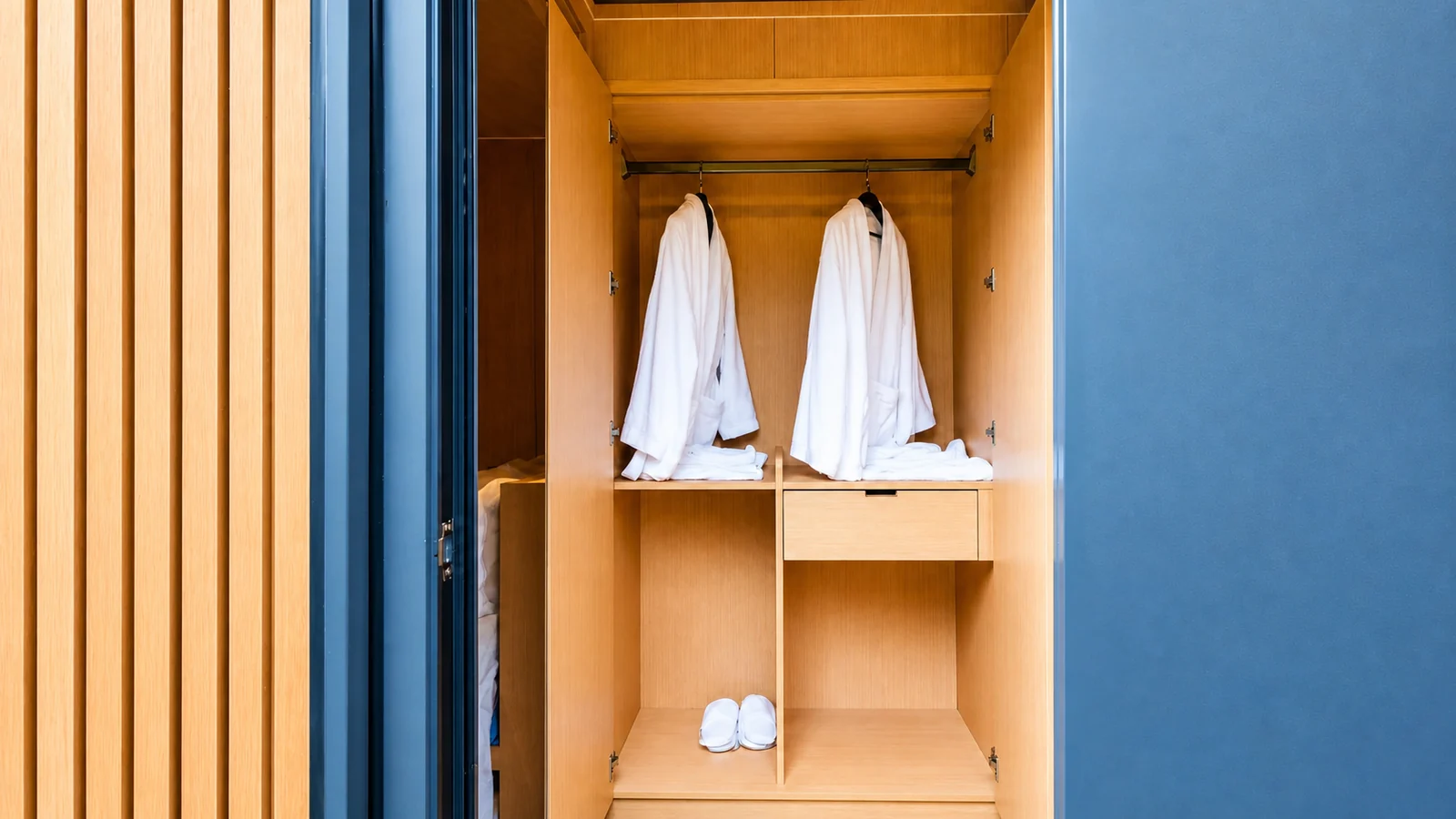 Open wardrobe in the timber clad capsule, two white robes and folded towels on timber shelves beside a fluted timber panel