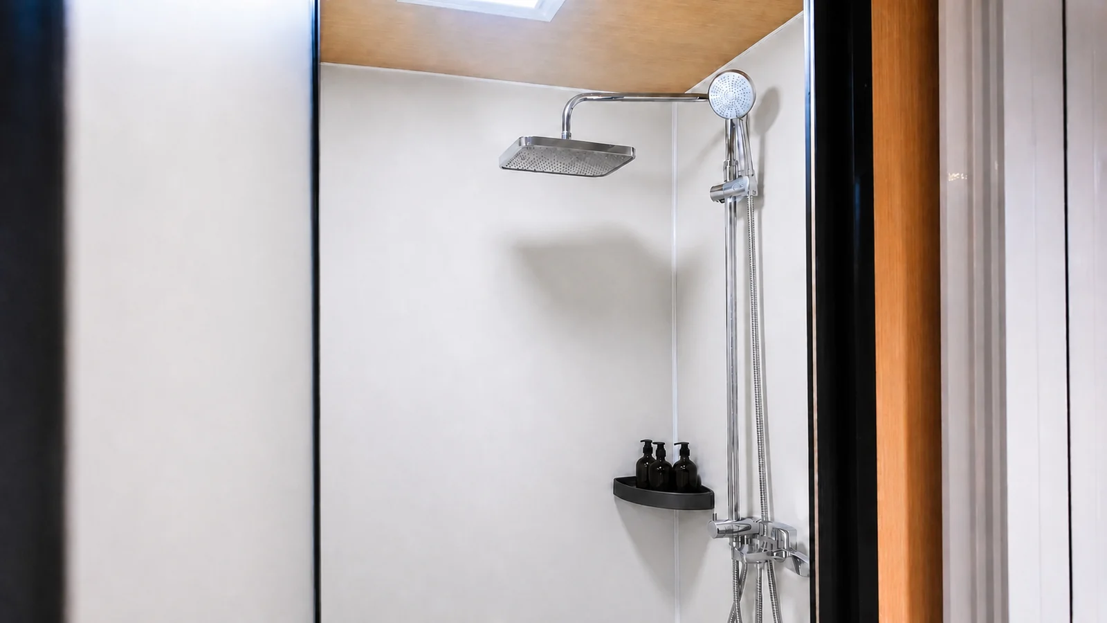 Shower in the timber clad capsule, chrome rain head and hand shower on a white wall with a black shelf holding bottles
