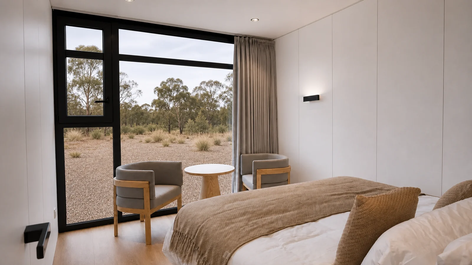 Bedroom in the 9.5 m timber clad capsule, made bed with a caramel throw and two armchairs by the full height glazed corner