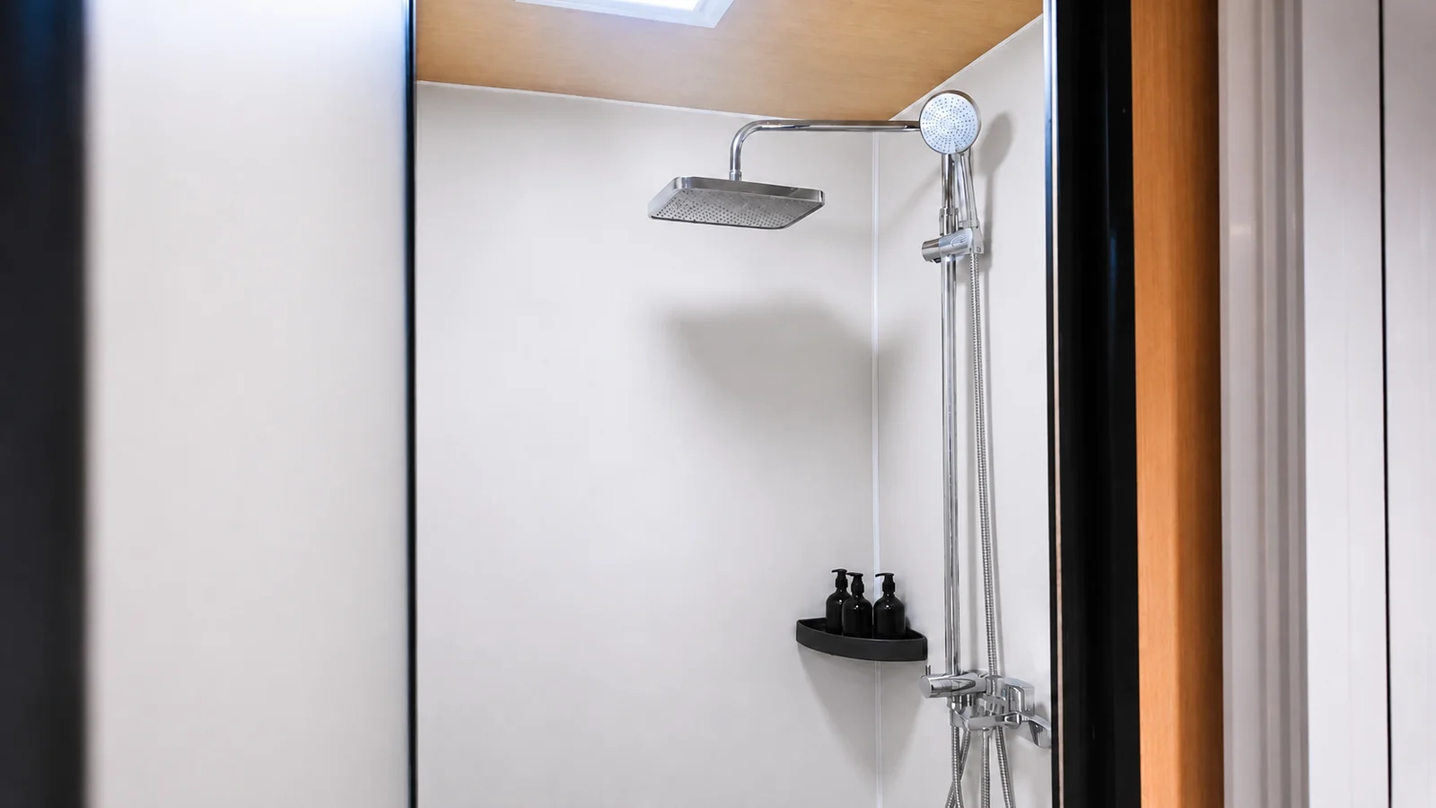 Shower in the 9.5 m timber clad capsule, chrome rain head and hand shower with a black shelf of toiletries on a white wall
