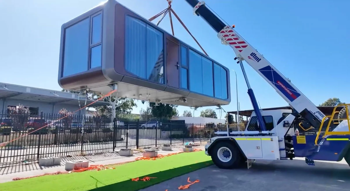 City crane lifting a capsule modular home clear of a black steel fence, with concrete pier footings and orange lifting slings laid out on the ground below