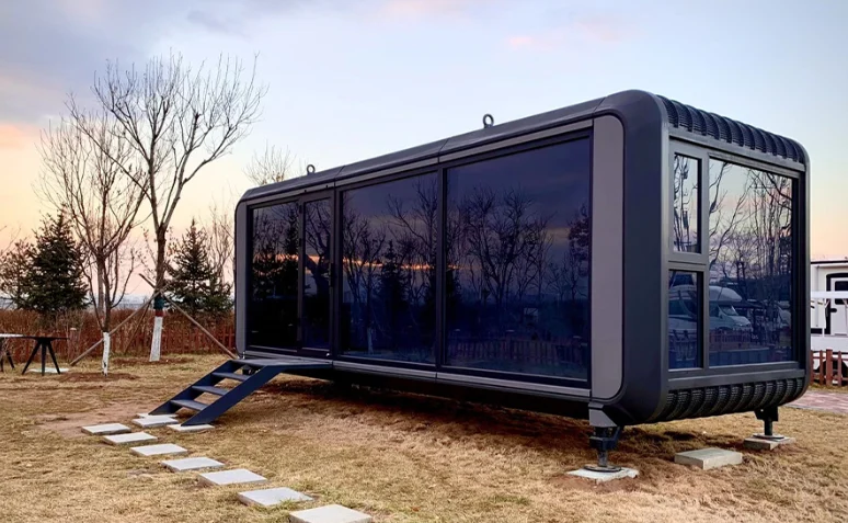 Dark capsule modular home levelled on adjustable feet over concrete pad footings at dusk, with steps running down to a paved path
