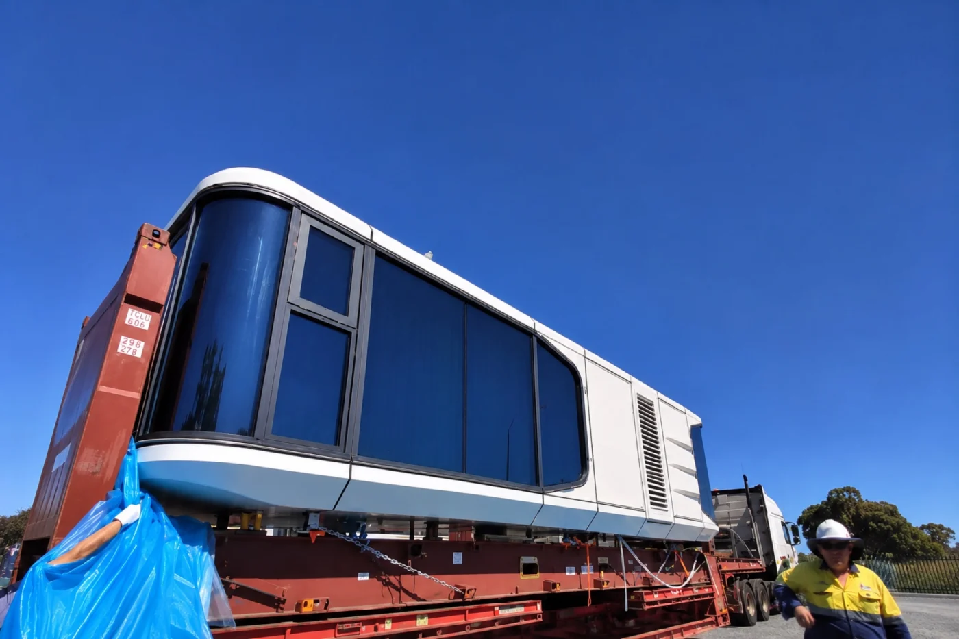 Capsule modular home strapped to a red low loader trailer behind a prime mover, with a worker in a hi-vis shirt and hard hat walking past under a clear blue sky